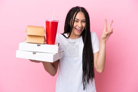 Téléchargez les photos : Adolescente avec des tresses tenant des pizzas et des hamburgers sur fond rose isolé souriant et montrant signe de victoire - en image libre de droit