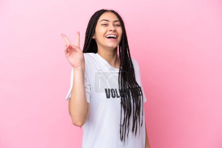 Téléchargez les photos : Adolescente bénévole avec des tresses isolées sur fond rose souriant et montrant le signe de la victoire - en image libre de droit