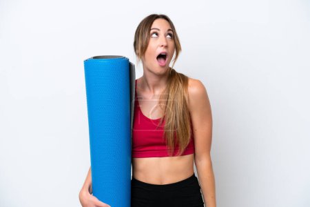 Téléchargez les photos : Jeune femme sportive allant à des cours de yoga tout en tenant un tapis isolé sur fond blanc levant les yeux et avec une expression surprise - en image libre de droit