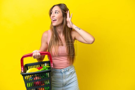 Photo for Young caucasian woman holding a shopping basket full of food isolated on yellow background listening to something by putting hand on the ear - Royalty Free Image