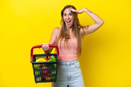 Photo for Young caucasian woman holding a shopping basket full of food isolated on yellow background with surprise expression - Royalty Free Image
