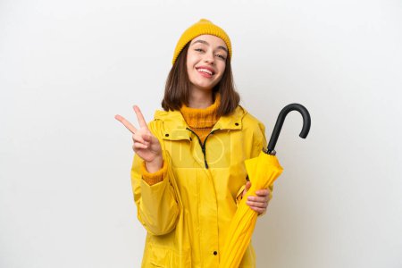Young Ukrainian woman with rainproof coat and umbrella isolated on white background smiling and showing victory sign