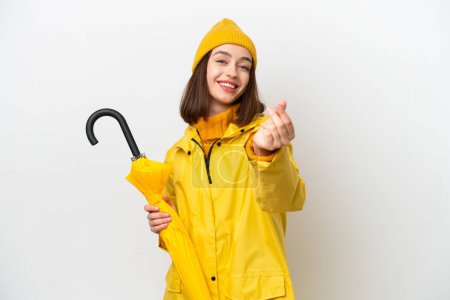 Junge Ukrainerin mit regendichtem Mantel und Regenschirm auf weißem Hintergrund macht Geld-Geste