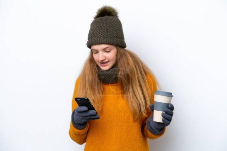 Téléchargez les photos : Adolescente russe portant une veste d'hiver isolée sur fond blanc tenant du café à emporter et un mobile - en image libre de droit