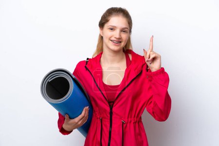 Foto de Adolescente chica yendo a clases de yoga mientras sostiene una estera aislada en bakcground blanco mostrando y levantando un dedo en señal de la mejor - Imagen libre de derechos