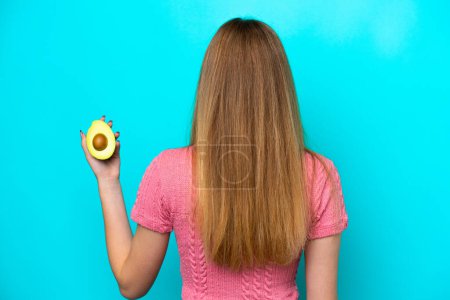 Photo for Teenager Russian girl holding an avocado isolated on blue background in back position - Royalty Free Image
