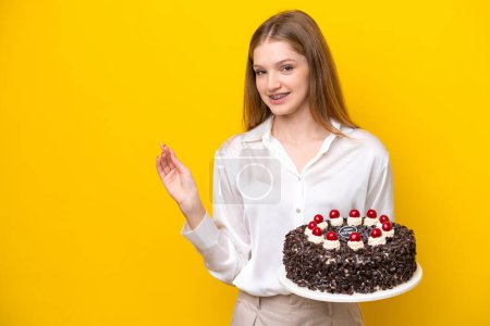 Foto de Adolescente rusa chica sosteniendo pastel de cumpleaños aislado sobre fondo amarillo apuntando hacia atrás - Imagen libre de derechos
