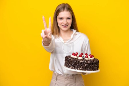 Foto de Adolescente rusa chica sosteniendo pastel de cumpleaños aislado sobre fondo amarillo sonriendo y mostrando signo de victoria - Imagen libre de derechos
