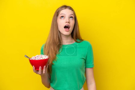 Téléchargez les photos : Adolescente russe tenant bol de céréales isolé sur fond jaune levant les yeux et avec expression surprise - en image libre de droit