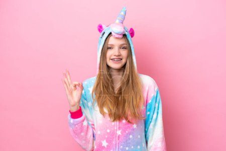 Foto de Adolescente chica rusa con pijama de unicornio aislado sobre fondo rosa mostrando signo de ok con los dedos - Imagen libre de derechos