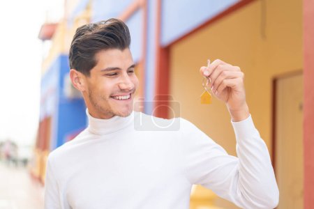 Téléchargez les photos : Jeune homme caucasien tenant les clés de la maison à l'extérieur avec expression heureuse - en image libre de droit