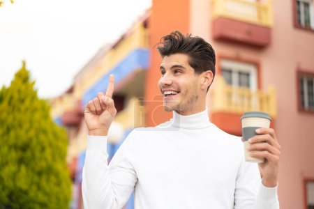 Foto de Joven hombre caucásico sosteniendo un café para llevar al aire libre con la intención de realizar la solución mientras levanta un dedo - Imagen libre de derechos