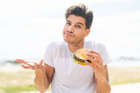 Young caucasian man holding a burger at outdoors making doubts gesture while lifting the shoulders