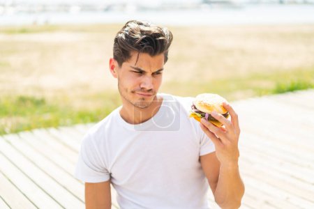 Photo for Young caucasian man holding a burger at outdoors with sad expression - Royalty Free Image