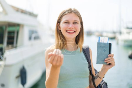 Téléchargez les photos : Jeune femme blonde tenant un passeport à l'extérieur invitant à venir avec la main. Heureux que tu sois venu - en image libre de droit