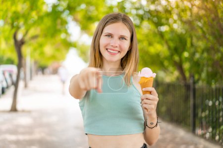 Téléchargez les photos : Jeune blonde jolie femme avec une crème glacée cornet à l'extérieur pointe du doigt vers vous avec une expression confiante - en image libre de droit