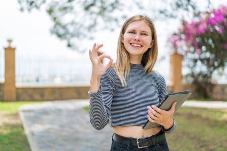 Foto de Mujer rubia joven sosteniendo una tableta al aire libre mostrando signo de ok con los dedos - Imagen libre de derechos