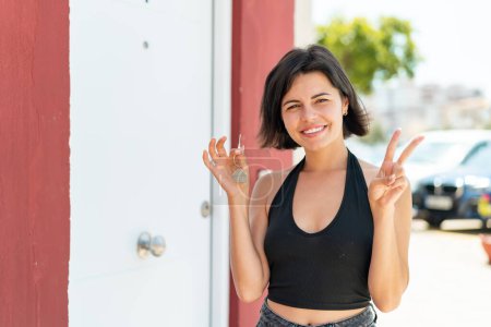 Téléchargez les photos : Jeune jolie bulgare femme tenant les clés de la maison à l'extérieur souriant et montrant signe de victoire - en image libre de droit