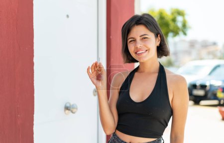 Foto de Joven bonita mujer búlgara sosteniendo las llaves de casa al aire libre sonriendo mucho - Imagen libre de derechos