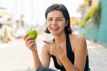 Téléchargez les photos : Jeune jolie femme bulgare à l'extérieur tenant une pomme avec une expression heureuse - en image libre de droit