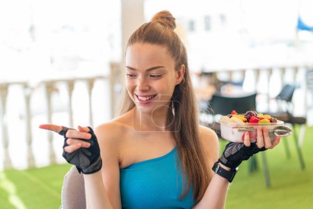 Téléchargez les photos : Jeune jolie fille sportive tenant un bol de fruits à l'extérieur pointant vers le côté pour présenter un produit - en image libre de droit