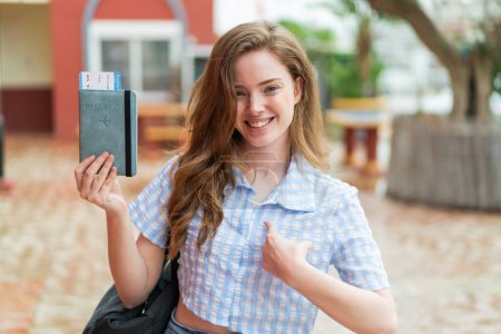 Téléchargez les photos : Jeune rousse femme tenant un passeport à l'extérieur avec expression faciale surprise - en image libre de droit
