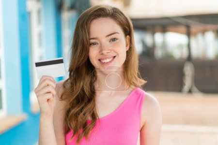Téléchargez les photos : Jeune rousse femme tenant une carte de crédit à l'extérieur souriant beaucoup - en image libre de droit