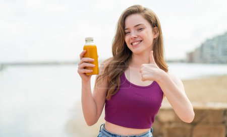 Téléchargez les photos : Jeune fille rousse tenant un jus d'orange à l'extérieur avec les pouces vers le haut parce que quelque chose de bon est arrivé - en image libre de droit