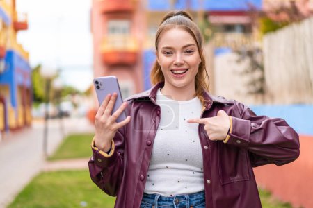 Téléchargez les photos : Jeune jolie fille utilisant téléphone mobile à l'extérieur avec expression faciale surprise - en image libre de droit