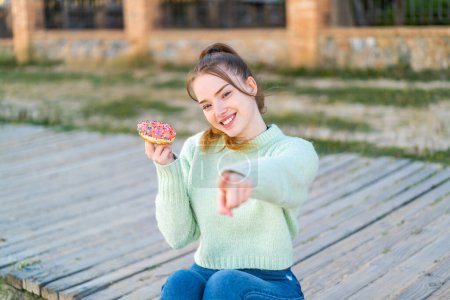 Foto de Joven chica bonita sosteniendo una dona al aire libre señala el dedo en usted con una expresión de confianza - Imagen libre de derechos