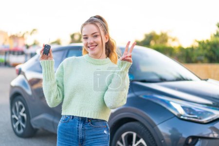 Foto de Joven chica bonita sosteniendo las llaves del coche al aire libre sonriendo y mostrando la señal de victoria - Imagen libre de derechos