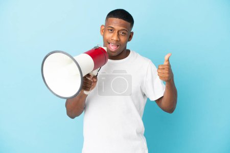 Téléchargez les photos : Jeune homme latin isolé sur fond bleu tenant un mégaphone avec pouce vers le haut - en image libre de droit