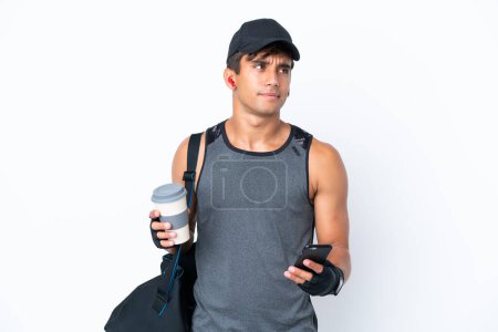 Téléchargez les photos : Jeune homme caucasien de sport avec sac de sport isolé sur fond blanc tenant du café à emporter et un mobile tout en pensant quelque chose - en image libre de droit