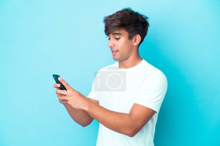 Téléchargez les photos : Jeune homme caucasien isolé sur fond bleu envoyer un message ou un e-mail avec le mobile - en image libre de droit