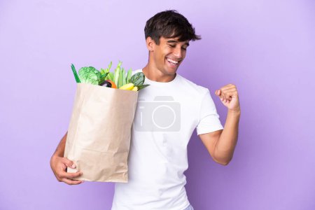 Photo for Young man holding a grocery shopping bag isolated on purple background celebrating a victory - Royalty Free Image