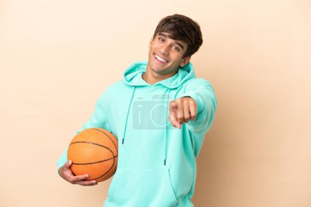 Téléchargez les photos : Beau jeune joueur de basket-ball isolé sur fond ocre pointant devant avec expression heureuse - en image libre de droit
