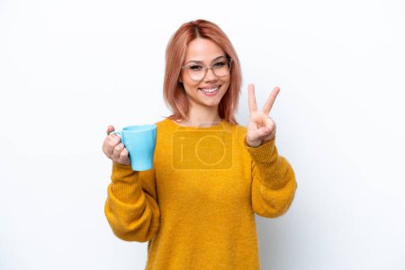 Foto de Joven chica rusa sosteniendo taza de café aislado sobre fondo blanco sonriendo y mostrando signo de victoria - Imagen libre de derechos