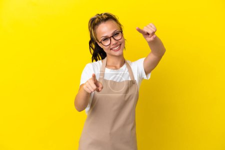 Photo for Restaurant waiter Russian girl isolated on yellow background points finger at you while smiling - Royalty Free Image