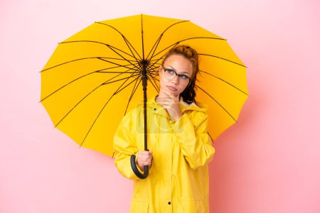Adolescente russe avec manteau imperméable et parapluie isolé sur fond rose ayant des doutes