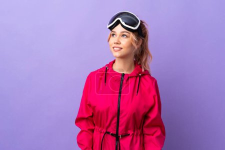 Téléchargez les photos : Skier adolescente avec des lunettes de snowboard sur fond violet isolé penser une idée tout en levant les yeux - en image libre de droit