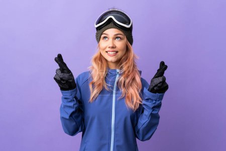 Foto de Esquiador adolescente con gafas de snowboard sobre fondo púrpura aislado con los dedos cruzando y deseando lo mejor - Imagen libre de derechos