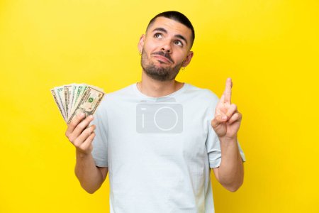 Téléchargez les photos : Jeune homme caucasien prenant beaucoup d'argent isolé sur fond jaune avec les doigts croisés et souhaitant le meilleur - en image libre de droit