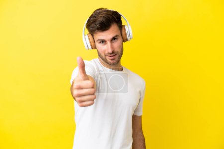 Foto de Joven hombre guapo caucásico aislado sobre fondo amarillo escuchando música y con el pulgar hacia arriba - Imagen libre de derechos