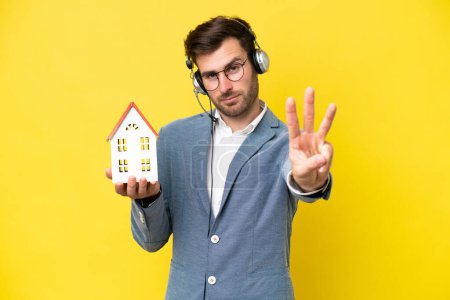 Foto de Joven hombre caucásico sosteniendo una casa aislada sobre fondo blanco feliz y contando tres con los dedos - Imagen libre de derechos