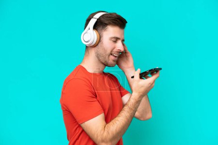Photo for Young caucasian man isolated on blue background listening music with a mobile and singing - Royalty Free Image