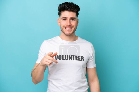 Young volunteer caucasian man isolated on blue background surprised and pointing front