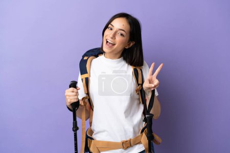 Téléchargez les photos : Jeune femme caucasienne avec sac à dos et bâtons de trekking isolés sur fond bleu souriant et montrant signe de victoire - en image libre de droit