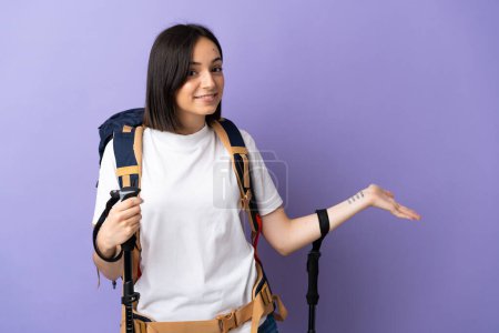 Foto de Mujer joven caucásica con mochila y bastones de trekking aislados sobre fondo azul extendiendo las manos a un lado para invitar a venir - Imagen libre de derechos