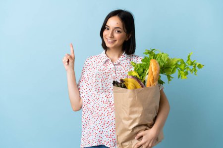 Foto de Mujer joven sosteniendo una bolsa de la compra de comestibles mostrando y levantando un dedo en señal de la mejor - Imagen libre de derechos