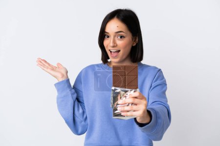 Mujer joven caucásica aislada sobre fondo blanco tomando una tableta de chocolate y sorprendida
